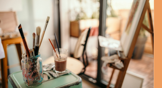 Photograph showing an easel set up in a person's kitchen in the background, with jars full of paintbrushes and trays of paint colours in the foreground